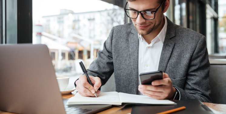 Homem branco de roupa formal e óculos de grau em um escritório na frente do notebook. Ele está fazendo anotações no caderno e segurando um celular.