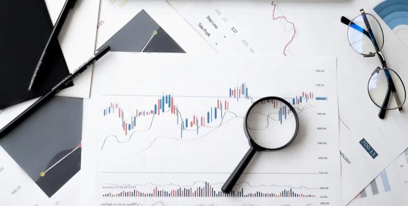 Análise de gráficos financeiros, relatório com dados econômicos, lupa, canetas, computador portátil e óculos em mesa de escritório, representando soluções de gestão tributária e consultoria financeira da Dootax.