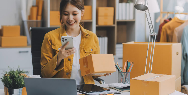 1. Mulher feliz usando celular em escritório de loja de comércio eletrônico com caixas de transporte, representando soluções de entrega e logística com Dootax.